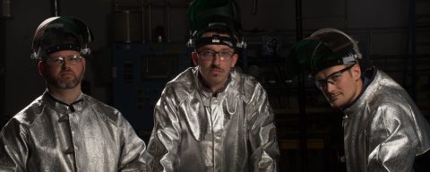 Three men, wearing glass-making protect suits, look into camera