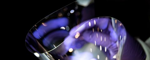 A pair of disembodied hands in purple gloves holding bendable glass in a stylized dark shot.